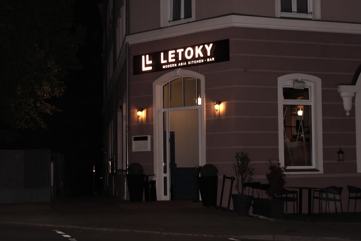 Das LETOKY am Abend von der Straßenecke aus: der beleuchtete Schriftzug über dem Eingang, davor die Terrasse mit Stühlen