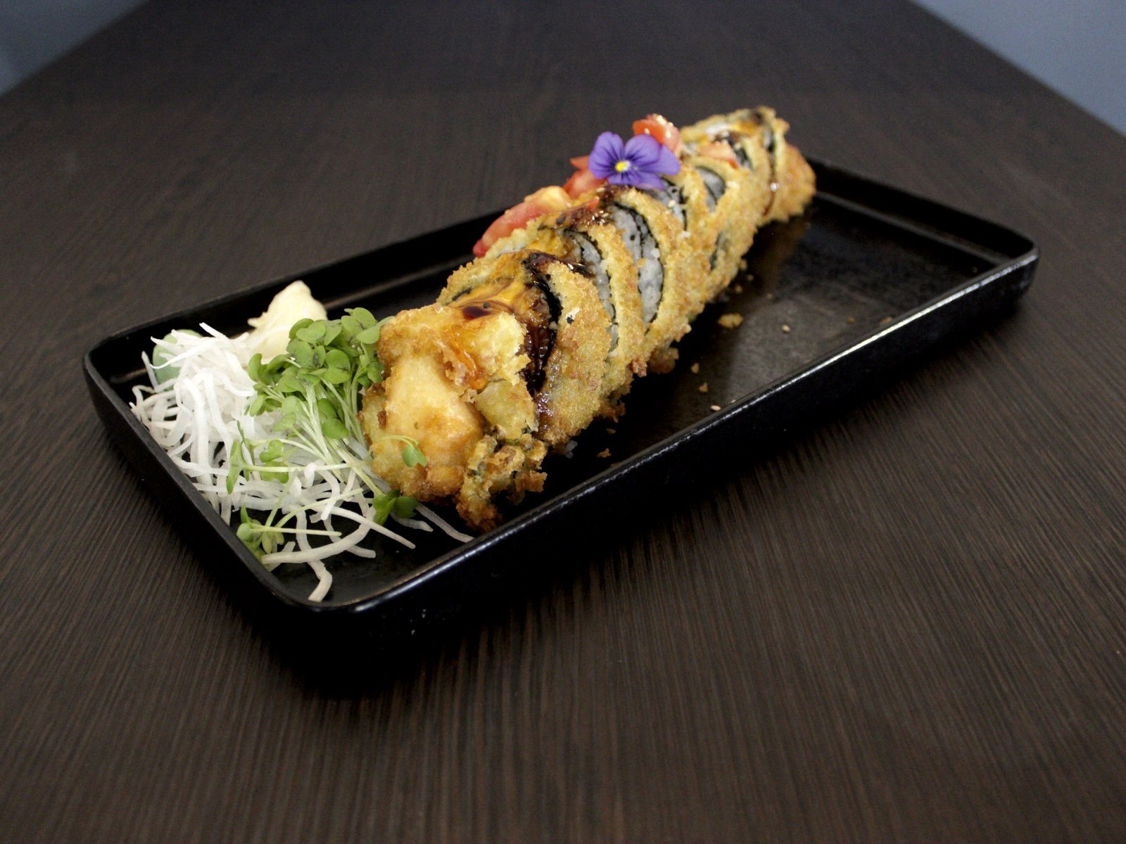 Frittierte Sushi-Rolle mit Sesam-Panade auf schwarzem Teller, dazu Rettich-Julienne, Kresse und eine essbare Blüte