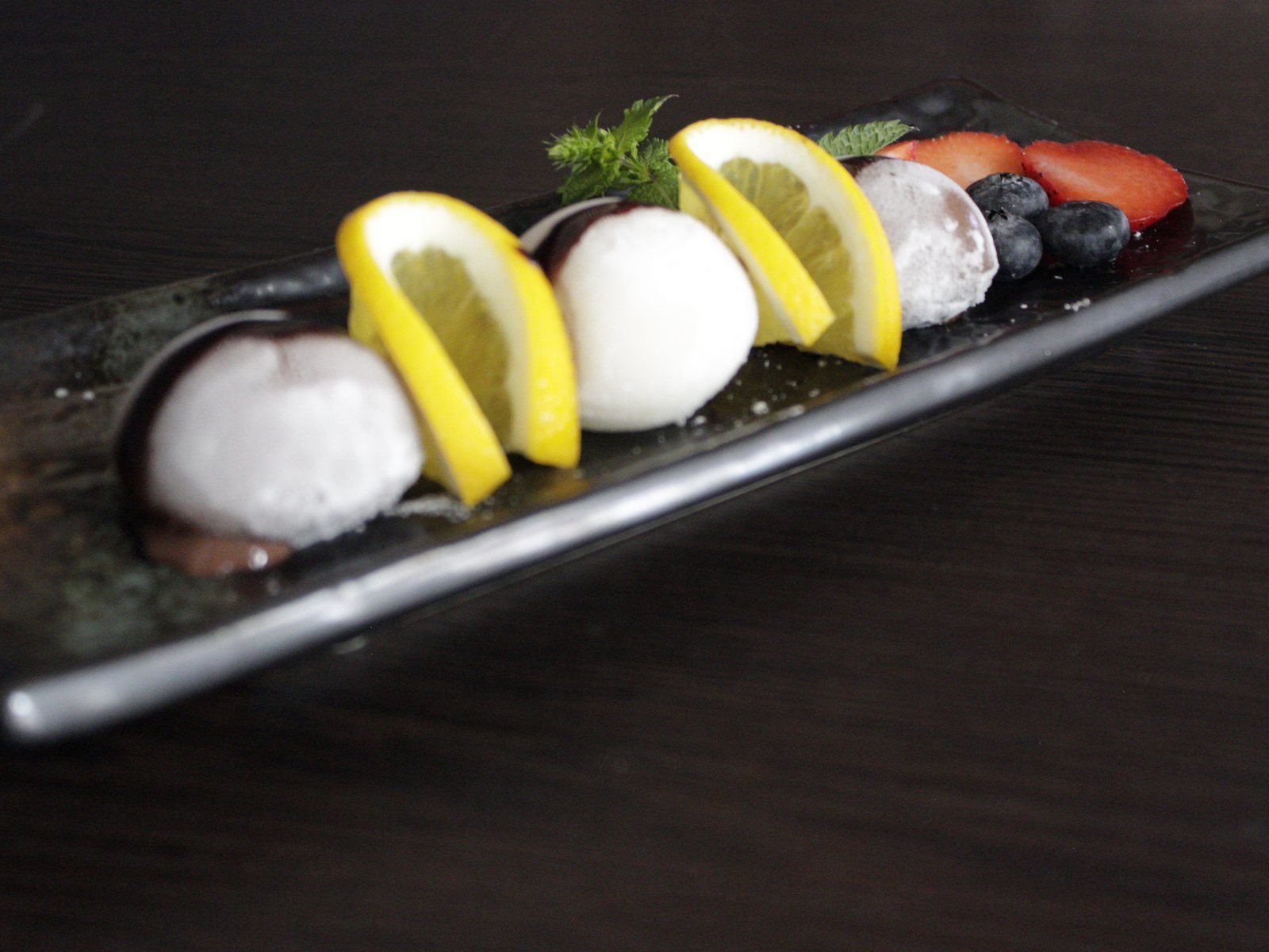 Längliche Platte mit drei Mochi-Eiskugeln, Schokoladensauce, Zitronenspalten, Beeren und Minze