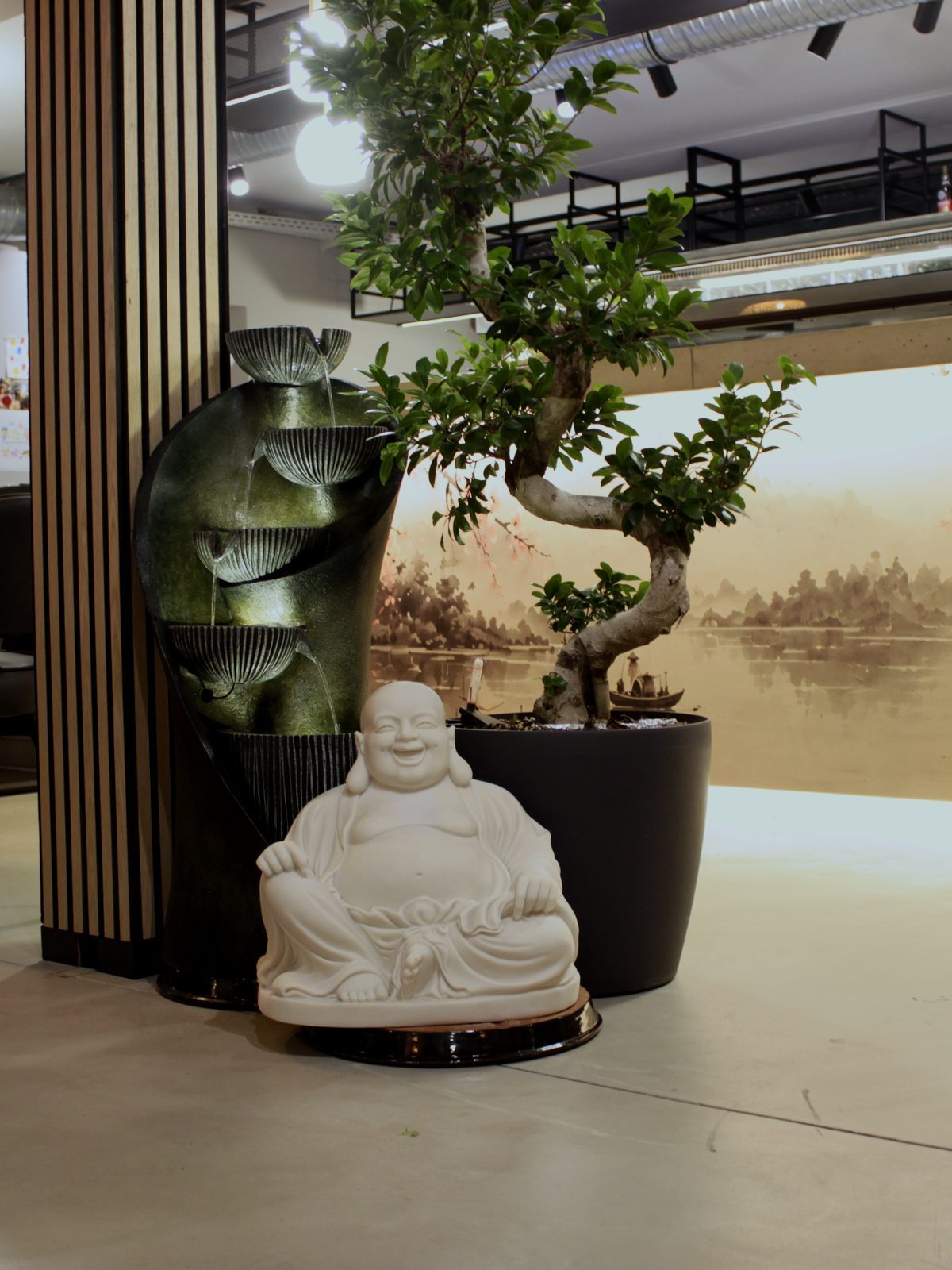 Weiße Buddha-Figur und ein Bonsai vor einem Wandbild im Tuschestil, daneben ein Zimmerbrunnen